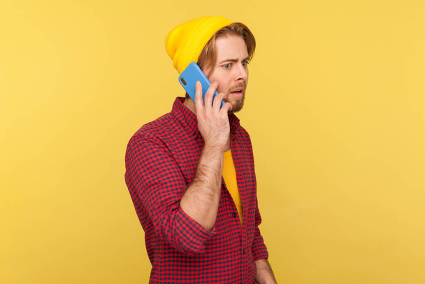 Portrait of hipster bearded guy in beanie hat and checkered shirt talking on phone, having serious mobile conversation, making important call. Indoor studio shot isolated on yellow background. - Photo, image