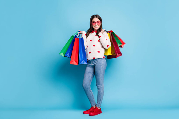 Full length photo of young woman dressed hearts print pullover spectacles holding shop bags isolated blue color background - Foto, Imagen