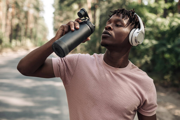 Sporty tired african american man , runner drinking water resting after workout, man relaxing after workout on city road in forest in nature - Φωτογραφία, εικόνα