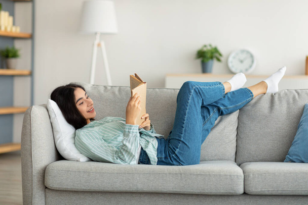 Zostań w domu. Millennial Indian kobieta leżąca na kanapie z otwartą książką w pomieszczeniach, ciesząc się spokojny weekend rano - Zdjęcie, obraz