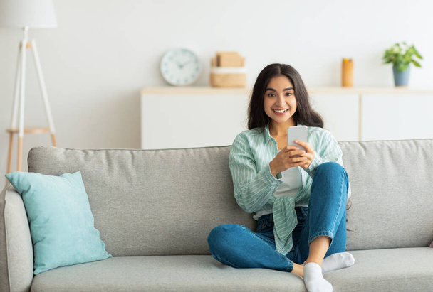 Очаровательная молодая индианка, сидящая на диване со смартфоном, проверяющая социальные сети, чатящаяся дома - Фото, изображение
