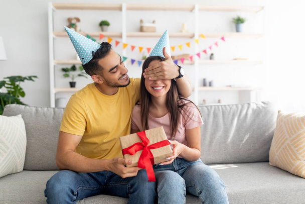 Hezký mladý Arab chlap zakrývá své přítelkyně oči, dělá narozeninové překvapení, dává jí zabalený dárek doma - Fotografie, Obrázek