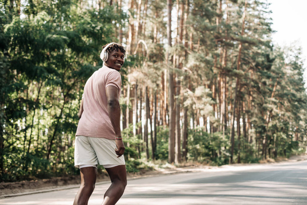 Corredor afroamericano feliz activo y saludable, escuchando música con auriculares, trotando al aire libre en la carretera en el bosque, vista lateral del joven atleta corriendo en la naturaleza - Foto, imagen