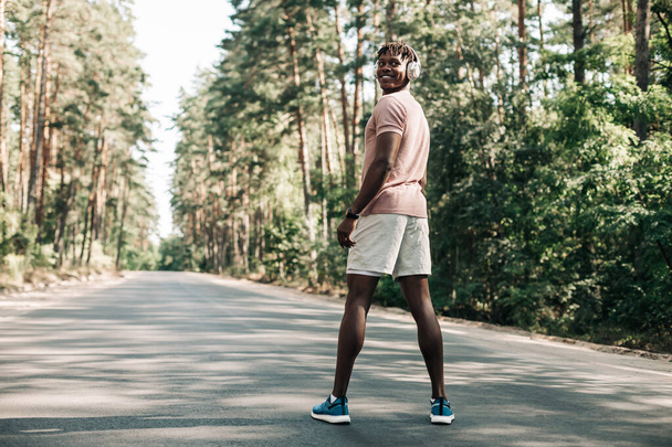 Μαύρος αθλητικός άνδρας, ντυμένος με αθλητικά ακούγοντας μουσική σε ακουστικά κατά τη διάρκεια της πρωινής γυμναστικής, Αφροαμερικάνος που γυμνάζεται σε εξωτερικούς χώρους και στέκεται στο δρόμο στο δάσος απολαμβάνοντας μουσική - Φωτογραφία, εικόνα