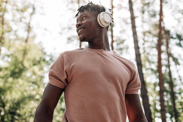 Black sporty man, dressed in sportswear listening to music on headphones during morning workout, African American man exercising in tropical park enjoying music - Fotó, kép