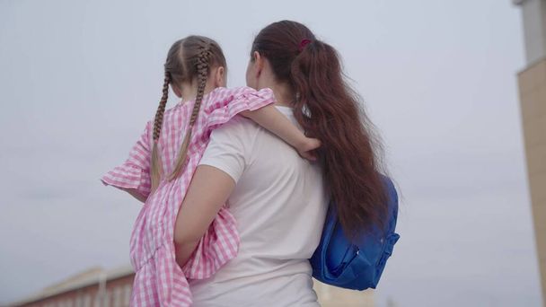 madre lleva a una pequeña hija con una mochila en sus brazos a la escuela, enseñando a un niño en la escuela primaria, una familia feliz, despedir a un niño con una bolsa de la escuela, adquirir conocimiento de la vida futura - Foto, Imagen