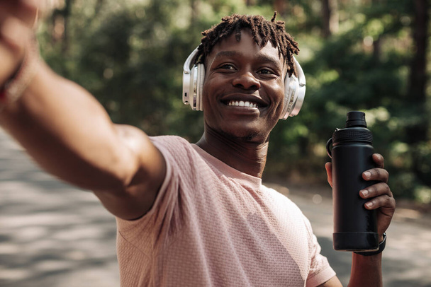 Portrait of smiling african american man wearing headphones taking selfie after jogging holding water bottle outdoors in park - Foto, Bild