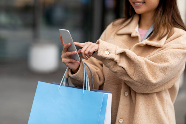 Primer plano de la mujer joven utilizando el teléfono inteligente, la celebración de bolsas de papel, compras en línea al aire libre - Foto, imagen