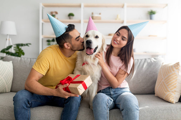 Young multinational couple in party hats celebrating dogs birthday, holding gift box, kissing their pet at home - Photo, Image