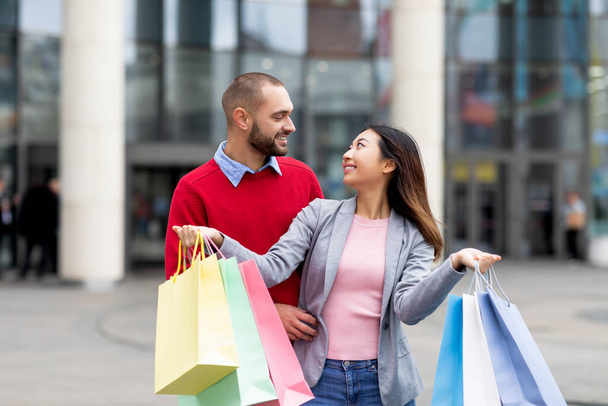 愛情のある異人種間のカップルがショッピングバッグとモールの近くに立って、お互いを見て、購入に満足 - 写真・画像