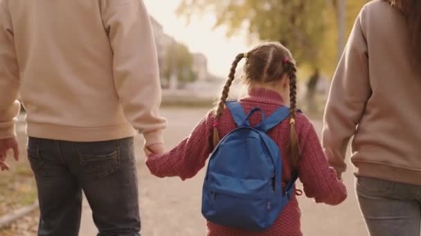 mother and father hold a little schoolgirl with a backpack by the hand, happy family, walk through schoolyard in fall, mom, dad and kid with bag over their shoulders, accompany child to first grade - Footage, Video