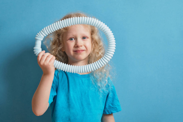 cute caucasian girl with curly blonde hair playing with pop it tube toy on blue background, trend toy for the development of fine motor skills of hands, anti stress crackling tube - Fotografie, Obrázek