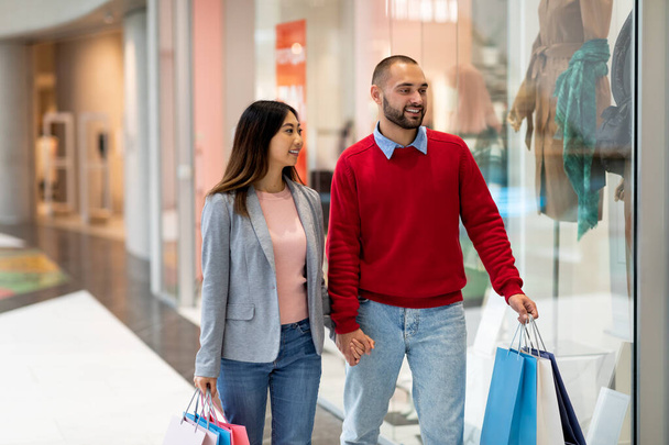 Loving interracial couple walking along store windows, holding hands, shopping for clothes at mall, copy space - Photo, Image