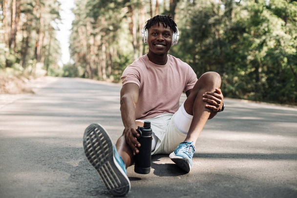 Happy african fitness man sitting relaxed on the road in the park, with a bottle of water after workout, sportsman taking a break after exercise outdoors in nature - Foto, Imagem