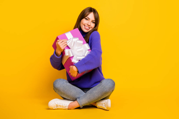 Photo of shiny sweet young lady dressed violet sweater sitting floor legs crossed hugging gift box isolated yellow color background - Fotoğraf, Görsel