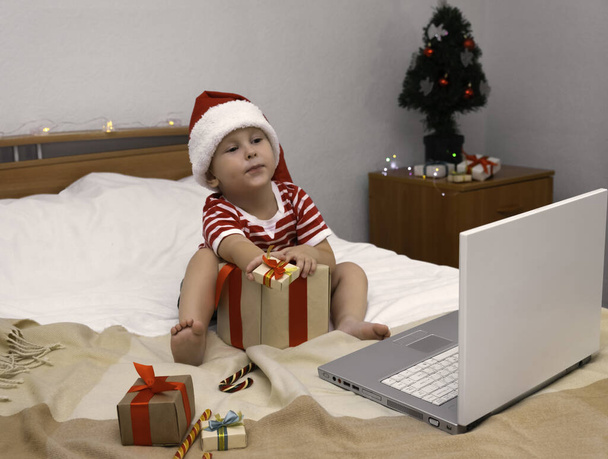 A child in a Christmas hat sits on the bed and talks via video link through a laptop, shows gifts. - Foto, Bild