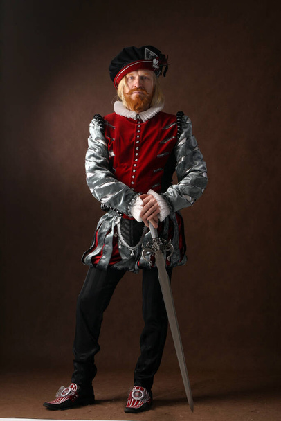 close-up portrait of a man of the Middle Ages with a beard and mustache in a suit isolated on a dark background with a sword standing - Photo, Image