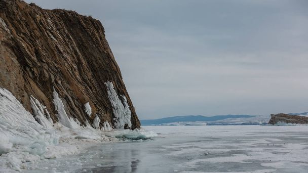 Ein schierer Granitfelsen ohne Vegetation erhebt sich auf einem zugefrorenen See. Die Basis ist eisig. Ein Gebirge vor wolkenverhangenem Himmel. Spiegelung auf dem Eis. Baikal - Foto, Bild