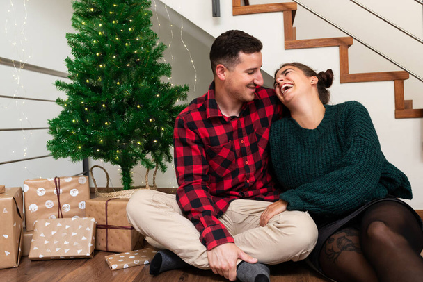 Junges verliebtes kaukasisches Paar lacht zusammen neben Weihnachtsbaum und einem Stapel Geschenke. - Foto, Bild