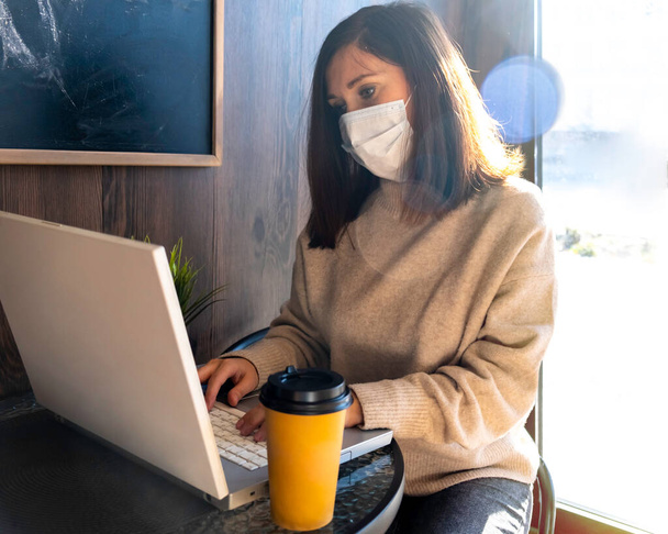 A girl in a medical mask in a cafe works at a laptop. Remote work, Study, pastime. - Фото, изображение