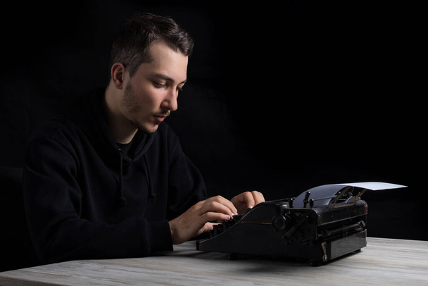 Jovem bonito homem de capuz preto está digitando em uma máquina de escrever, em um fundo preto em chave baixa - Foto, Imagem