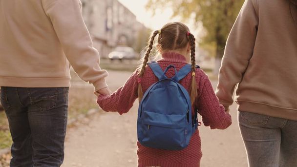 moeder en vader houden een klein schoolmeisje met een rugzak bij de hand, gelukkig gezin, lopen in de herfst door het schoolplein, moeder, vader en kind met tas over hun schouders, begeleiden kind naar de eerste klas - Foto, afbeelding