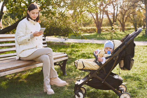 Beautiful young caucasian woman sitting on a park bench with her little son in a stroller, texting on a mobile phone and smiling. Full view. - Foto, Imagen