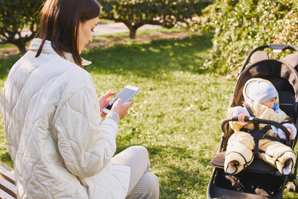 Modern young mother, sitting on a bench in an autumn park with her little son in a stroller, holding a smartphone in her hand, exchanging messages, working remotely. Mom concept on maternity leave. - Foto, immagini
