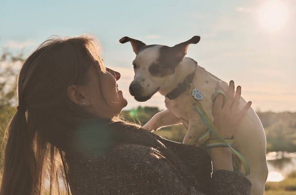 Sonbahar kıyafetli genç bir kadın köpeğini tutuyor ve onu kucaklıyor. Sevgi dolu evcil hayvan. Parkta ışıklandırılmış. Sahibi ve Jack Russell Terrier 'ın yan görüntüsü - Fotoğraf, Görsel