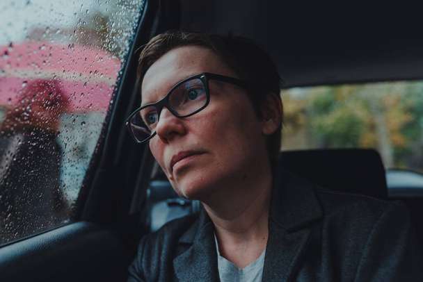 Worried businesswoman with eyeglasses waiting in the car and looking out the window during rain, selective focus - Foto, Imagen