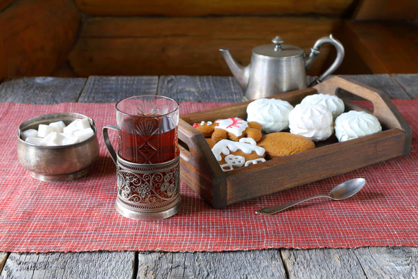 Un vaso de té negro está en la mesa de madera entre utensilios de plata y una bandeja con galletas de jengibre y malvaviscos.  - Foto, imagen