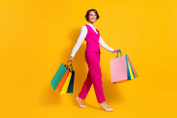 Photo of sweet shiny young woman dressed pink suit holding shopping bags walking isolated yellow color background - Foto, Imagen