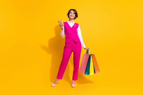 Φωτογραφία από ονειρική γλυκιά νεαρή γυναίκα ντυμένη ροζ κοστούμι κρατώντας αγοραστές σύγχρονο gadget αναζητούν κενό χώρο απομονωμένο κίτρινο χρώμα φόντο - Φωτογραφία, εικόνα