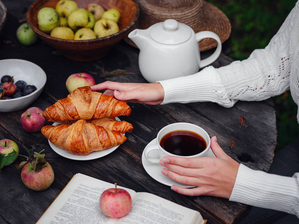 podzimní byt ležel tmavý dřevěný stůl na zahradě nebo na terase ženské ruce s kávou, jablky, croissanty, švestkami a knihou. myšlenka a koncepce teplého podzimu, útulný životní styl a udržitelný jednoduchý život - Fotografie, Obrázek