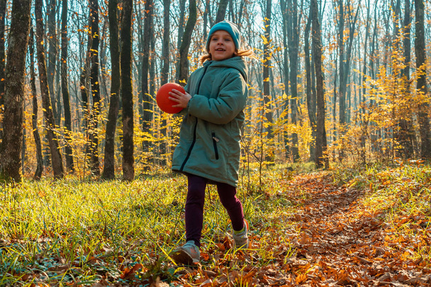 A child girl in a jacket and cap with a ball in her hands in an autumn forest. In the background is green grass, yellow leaves and tree trunks. The concept of a happy childhood - Fotografie, Obrázek