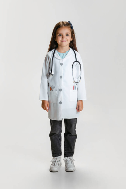 Full-length portrait of cute beautiful little girl, child in image of doctor wearing white lab coat posing isolated on white studio background - Foto, Imagem