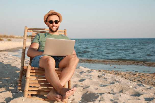 Man working with laptop in deck chair on beach. Space for text - 写真・画像