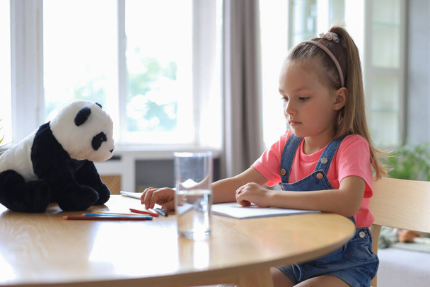 Smilling happy girl sitting in the table with a toy panda bear near to her enjoying creative activity, drawing pencils coloring pictures in albums - Valokuva, kuva