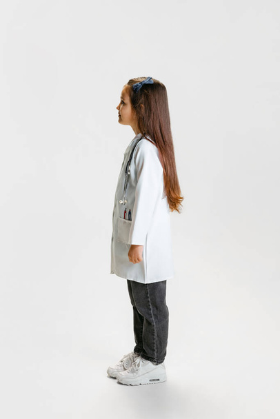 Side view. Cute beautiful little girl, child in image of doctor wearing white lab coat posing isolated on white studio background - 写真・画像