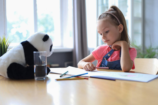 Smilling happy girl sitting in the table with a toy panda bear near to her enjoying creative activity, drawing pencils coloring pictures in albums - Φωτογραφία, εικόνα
