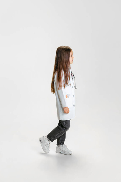 Side view. Cute beautiful little girl, child in image of doctor wearing white lab coat posing isolated on white studio background - Photo, Image
