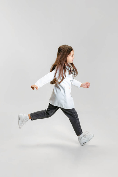 Full-length portrait of cute beautiful little girl, child in image of doctor wearing white lab coat running isolated on white studio background - Foto, immagini