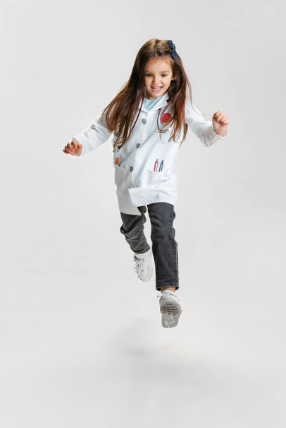Full-length portrait of cute beautiful little girl, child in image of doctor wearing white lab coat running isolated on white studio background - 写真・画像