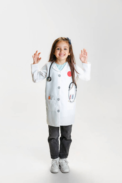 Full-length portrait of cute beautiful little girl, child in image of doctor wearing white lab coat posing isolated on white studio background - Foto, Imagem