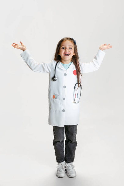 Preschool beautiful girl, child in image of doctor wearing white lab coat posing isolated on white studio background - Fotó, kép