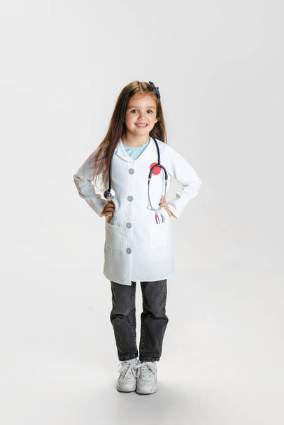 Preschool beautiful girl, child in image of doctor wearing white lab coat posing isolated on white studio background - Foto, afbeelding