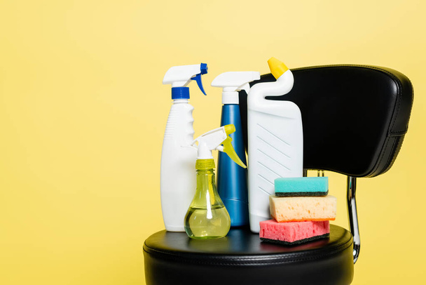 Flaschen mit Waschmittel und sauberen Schwämmen auf schwarzem Stuhl isoliert auf gelb - Foto, Bild