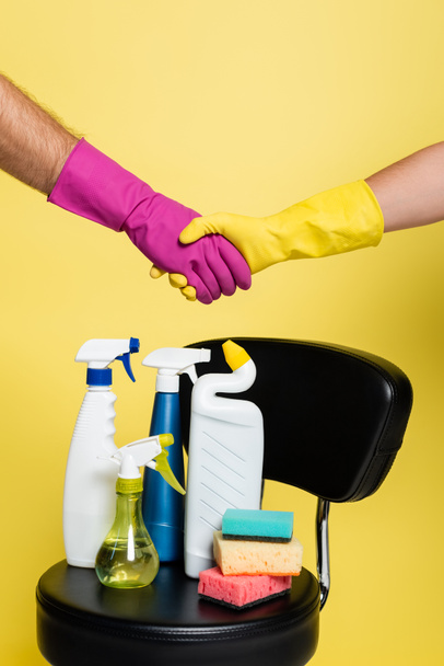 bijgesneden zicht van reinigers schudden handen in de buurt van het bereiken van flessen met wasmiddel en schone sponzen op stoel geïsoleerd op geel - Foto, afbeelding