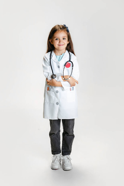 Portrait of cute beautiful girl, child in image of doctor wearing white lab coat posing isolated on white studio background - 写真・画像