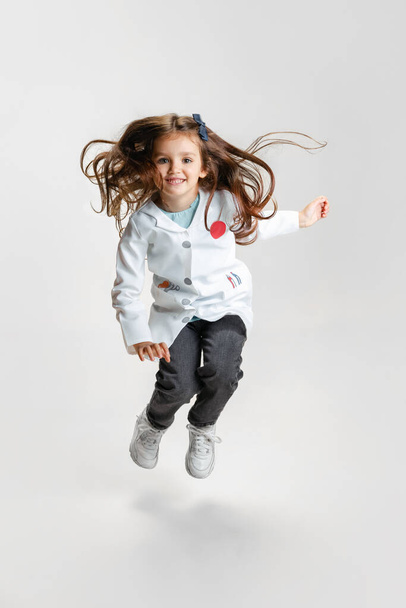 Full-length portrait of cute beautiful little girl, child in image of doctor wearing white lab coat jumping isolated on white studio background - Foto, Imagen